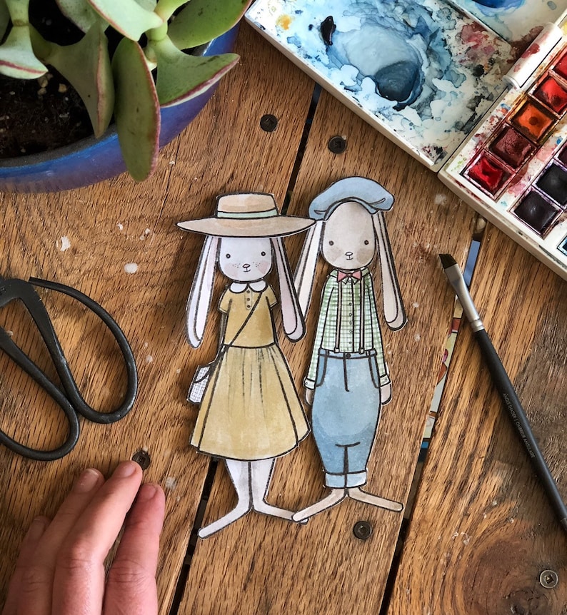 May include: Two paper dolls of bunnies, one wearing a straw hat and a yellow dress, the other wearing a blue hat and a green and white plaid shirt and blue pants.