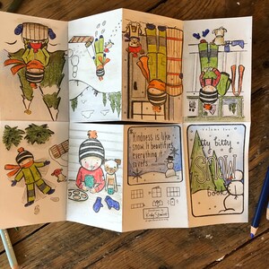 May include: A folded paper booklet with hand-drawn illustrations of winter scenes. The booklet features images of children playing in the snow, a snowman, and the text "Itty Bitty Snow Book". Colored pencils are scattered around the booklet.