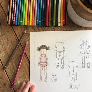 Puede incluir: Un conjunto de muñecas de papel en blanco y negro con una niña llamada Evie. El conjunto incluye una camiseta rosa, un vestido azul, una camisa a rayas blancas y negras y un mono blanco y negro. El conjunto también incluye un perro pequeño.