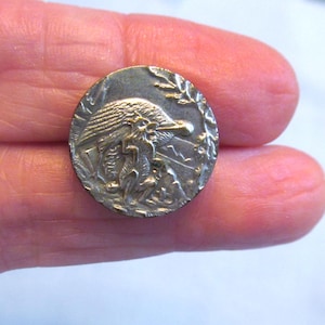 May include: A circular metallic button featuring a raised relief design of a fox standing on its hind legs in a forest setting. The button has a dark, antiqued silver-tone finish with detailed engravings of foliage and a small animal figure.