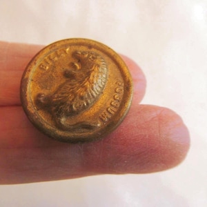 May include: A round, antique brass button with a detailed embossed design. The button features a possum image and the text "Billy Possum" around the edge. The button is held by a person's fingers.