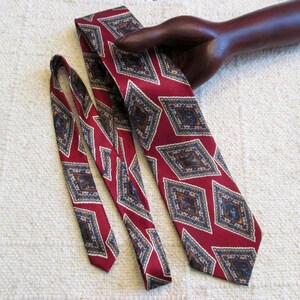 May include: A burgundy tie with a repeating diamond pattern in navy, tan, and cream. The tie is displayed with a matching tie and a wooden hand prop.