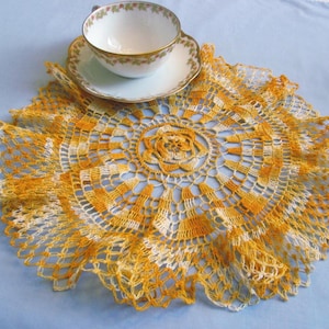 May include: A vintage teacup and saucer set atop a handmade, round doily. The doily features a detailed crochet pattern in shades of yellow and white, with a scalloped edge. The teacup and saucer are white with floral detailing and gold trim.