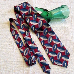 May include: A patterned necktie with a repeating oval design in red, blue, and white. The tie is lying on a textured, off-white surface. A green glass bottle is in the background.