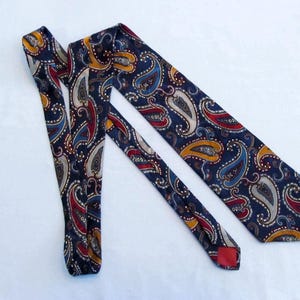 May include: A navy blue paisley tie with a repeating pattern of teardrop-shaped designs in red, yellow, blue, and white. The tie is made of a smooth fabric and is displayed on a white surface.