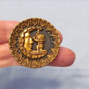 May include: A round, metallic gold-toned button featuring a raised relief design of a crescent moon with a human face and a small figure kneeling before it. The outer border is decorated with a repeating leaf-like pattern on a dark background.