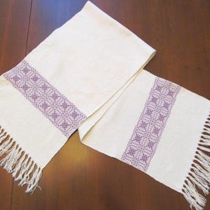 May include: A rectangular off-white table runner featuring two wide bands of intricate purple geometric patterns. The fabric has a textured weave and both ends of the runner are finished with long, loose white fringe tassels.