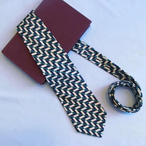 May include: A patterned tie with a geometric design in navy blue, cream, and dark green, draped over a burgundy-colored object. The tie is neatly coiled at the end, showcasing the repeating pattern. The background is a light blue surface.