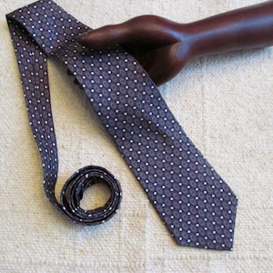 May include: A navy blue tie with a repeating white dot pattern and small red accents. The tie is displayed on a neutral-colored surface, with a portion of the tie coiled. The tie is held by a wooden hand.