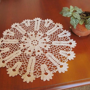 May include: A cream-colored, handmade crochet doily with a floral design, resting on a wooden table. The doily features a central flower pattern surrounded by intricate openwork and scalloped edges. A small potted plant with green and white leaves sits nearby.