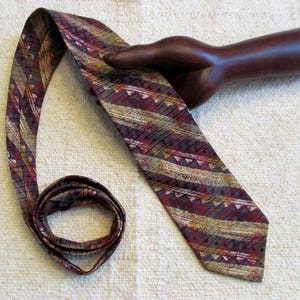 May include: A patterned necktie with a burgundy, gold, and gray design. The tie features a repeating pattern of stripes and triangles. The tie is displayed on a wooden hand sculpture.