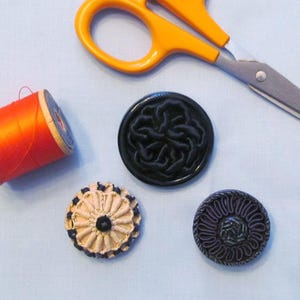 May include: A spool of bright orange thread, a pair of orange-handled metal scissors, and three circular buttons. One button is black with a raised knot pattern, one is beige with a pleated design, and one is black with a radial corded pattern.