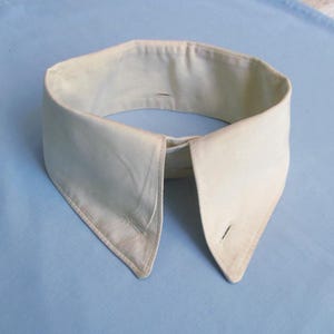 May include: An off-white, vintage detachable collar with pointed tips and buttonholes. The collar is made of a soft fabric and is set against a light blue background. A classic accessory.
