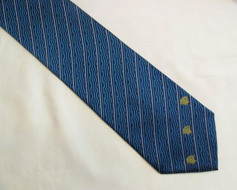 Puede incluir: Una corbata azul con un patr&oacute;n repetitivo de rayas verticales en azul oscuro y finas l&iacute;neas rosa claro. La parte inferior de la corbata muestra tres peque&ntilde;os emblemas bordados de color dorado dispuestos en una l&iacute;nea vertical cerca del borde.