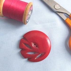 May include: A red, carved button with a floral design, a spool of pink thread, and orange-handled scissors on a light blue fabric. The button has two small holes for sewing.