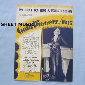 May include: Vintage sheet music cover for "Gold Diggers of 1933" featuring a woman in a glamorous gown. The title is in large yellow text, with the names of the stars and songwriters listed below. The text "I'VE GOT TO SING A TORCH SONG" is at the top.