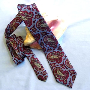 May include: A patterned necktie with a light blue background and a repeating paisley design in red and gold. The tie is lying on a white surface with a seashell in the background.
