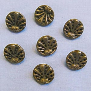 May include: A set of seven round, antique brass-colored buttons, each featuring a raised, symmetrical fan-like pattern with radiating lines and small circular cutouts. The buttons have a metallic finish and a distinct, textured relief design.