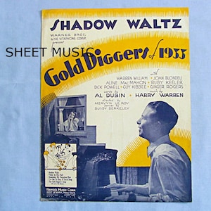 May include: A vintage sheet music cover for the song "Shadow Waltz" from the 1933 musical "Gold Diggers of 1933". The cover features a yellow and blue color scheme with a photo of Warren William playing the piano. The text on the cover includes the names of the cast members and the songwriters.