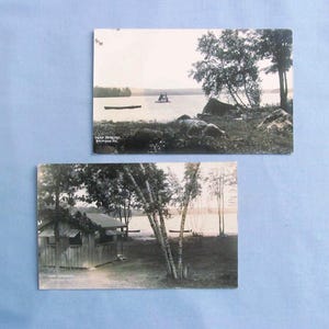 May include: Two vintage black and white postcards. One shows a lake scene with a boat and trees. The other depicts a small wooden cabin near a lake with trees. Both cards have text at the bottom.