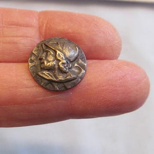 May include: A small, round, antique-style button with a detailed relief of a man's profile wearing a helmet. The button is made of a bronze-colored metal and has a slightly worn appearance, suggesting age and use. The design is intricate and well-preserved.