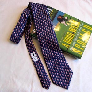 May include: Navy blue tie with a repeating pattern of pink and white shapes. The tie is draped over a book titled "A Good Walk Spoiled" with a green cover. The tie has a small tag with text.
