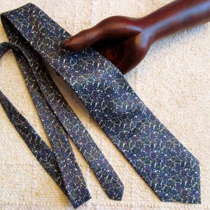 May include: A navy blue tie with a repeating floral pattern in shades of green, purple, and white. The tie is displayed on a cream-colored textured surface.