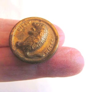 May include: A round, metallic, bronze-toned button featuring a raised relief design of an opossum in the center. The text "BILLY" is embossed along the upper edge, and the word "POSSUM" is embossed along the lower edge of the button surface.