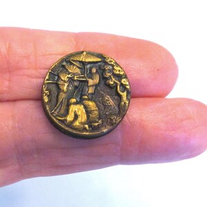 May include: A round, metallic button with a dark, aged brass finish. The surface features a raised, detailed relief depicting figures in traditional attire, one holding an umbrella, alongside a tree. The design is textured with dark recessed areas.