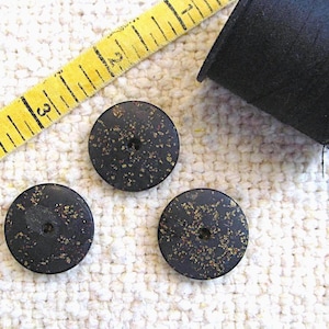 May include: Three round black buttons with gold-colored flecks, each featuring a single center hole. A yellow measuring tape shows the buttons are approximately 0.75 inches in diameter, positioned next to a spool of black sewing thread.