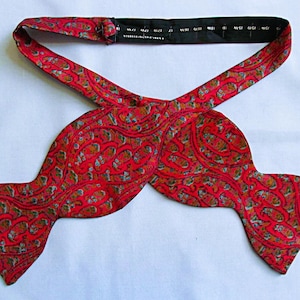 May include: A red bow tie featuring a repeating paisley pattern in shades of blue, green, and gold. The bow tie has an adjustable black neck strap with printed size markings ranging from 15 to 19 inches, including a metal hook and eye closure.