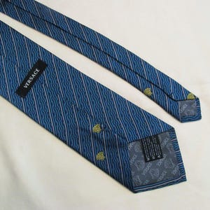 Puede incluir: Una corbata azul con un patr&oacute;n de rayas diagonales y peque&ntilde;os iconos amarillos de la cabeza de Medusa. La corbata incluye una etiqueta negra con el texto blanco VERSACE y un forro gris estampado con el logotipo de Versace y motivos de Medusa.