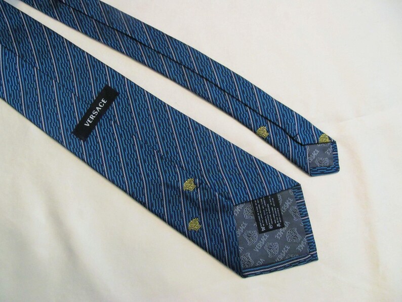 Puede incluir: Una corbata azul con un patr&oacute;n de rayas diagonales y peque&ntilde;os iconos amarillos de la cabeza de Medusa. La corbata incluye una etiqueta negra con el texto blanco VERSACE y un forro gris estampado con el logotipo de Versace y motivos de Medusa.