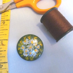 May include: A collection of sewing supplies, including a green button with a floral design, a spool of brown thread, yellow scissors, and a yellow measuring tape. The button features white and yellow flowers.
