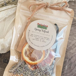 May include: A clear plastic bag filled with dried orange slices, cinnamon sticks, lavender, rosemary, and vanilla beans. The bag is tied with twine and has a label that reads "Spring Refresh Stove Top Simmer Pot Kit". The label also includes instructions for using the ingredients to make a simmer pot.