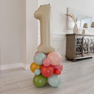 May include: A balloon arrangement for a first birthday celebration. A large beige number one balloon sits atop a cluster of colorful balloons in shades of pink, red, green, yellow, and blue. The balloons are arranged in a decorative display.