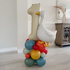 May include: A balloon arrangement for a first birthday celebration. It features a large beige number one balloon, a white goose balloon with yellow accents, and a cluster of colorful balloons in red, blue, yellow, and green.