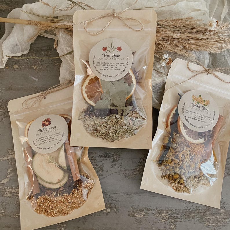 Peut inclure: Trois sachets transparents remplis d'herbes et d'&eacute;pices s&eacute;ch&eacute;es, chacun scell&eacute; avec de la ficelle. Les sachets ont des &eacute;tiquettes circulaires avec du texte. L'une des &eacute;tiquettes indique "Forest Spice", une autre "Fall Harvest" et la troisi&egrave;me "Golden Glow". Les sachets contiennent des tranches d'orange s&eacute;ch&eacute;es, des b&acirc;tons de cannelle et d'autres plantes.