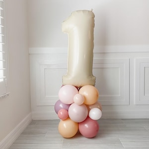 May include: A large, cream-colored number one balloon sits atop a cluster of pastel-colored balloons. The balloons are in shades of pink, peach, and lavender, creating a festive display. The arrangement is likely for a first birthday celebration.