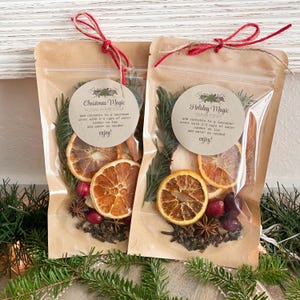Puede incluir: Dos bolsas de pl&aacute;stico transparente con respaldo de papel marr&oacute;n, cada una contiene un kit de olla de cocci&oacute;n a fuego lento para las fiestas. Las bolsas est&aacute;n atadas con hilo rojo y marr&oacute;n. Los kits incluyen rodajas de naranja secas, palitos de canela, an&iacute;s estrellado, clavos y ar&aacute;ndanos. Las bolsas tienen etiquetas que dicen "Christmas Magic Holiday Simmer Pot Kit" y "Holiday Magic Simmer Pot Kit".
