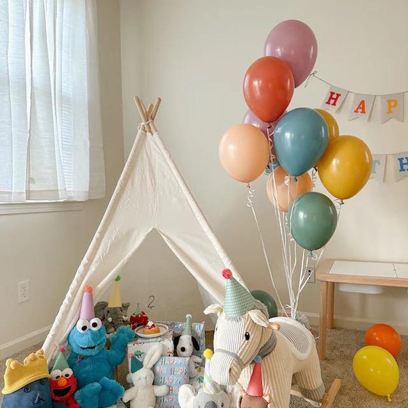 May include: A white teepee tent with a rocking horse and stuffed animals inside. Colorful balloons with the words "Happy Birthday" are in the background.