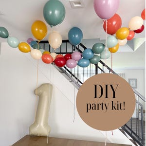 May include: A DIY party kit featuring a collection of latex balloons in muted shades of sage green, mustard yellow, dusty rose, terracotta, and slate blue, along with a large, beige, number one-shaped foil balloon and a circular graphic overlay.