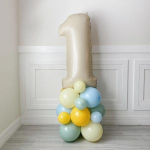 May include: A large beige number one balloon with a cluster of pastel blue, yellow, and cream balloons at the base.