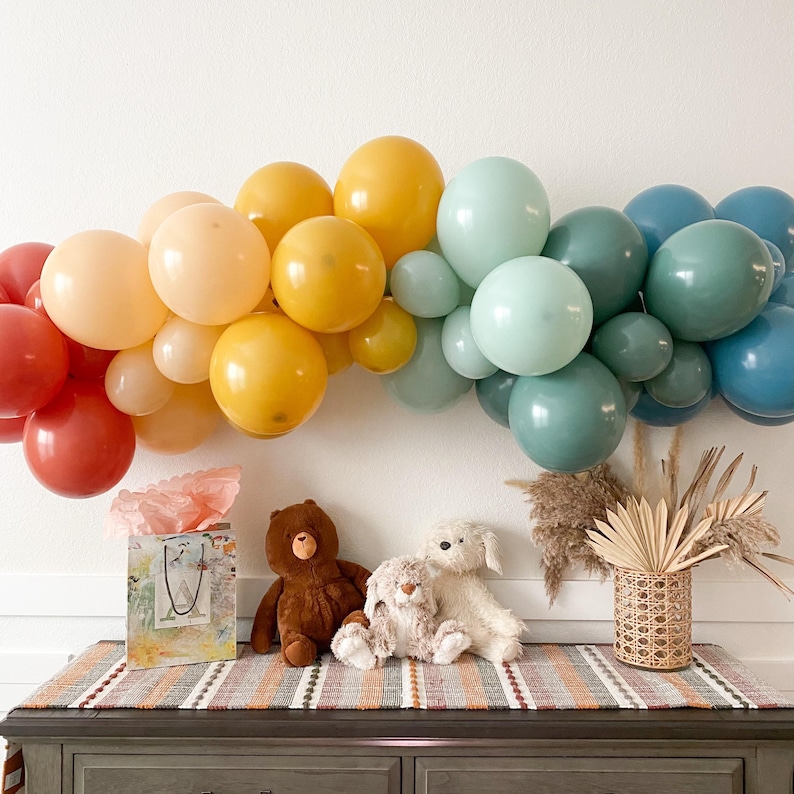 May include: A rainbow balloon arch made of red, orange, yellow, green, and blue balloons. Three stuffed toys, a brown bear, a white dog, and a brown bunny, are sitting on a striped rug in front of the balloon arch. A wicker basket with dried flowers is on the right side of the image.