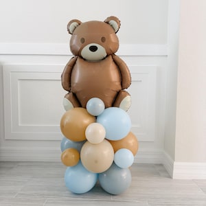 May include: A brown teddy bear balloon sits atop a cluster of balloons in shades of blue, tan, and gold. The bear has a light brown face and dark brown eyes. This balloon arrangement is suitable for a baby shower or birthday party.