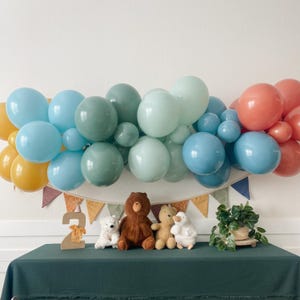 May include: A green, blue, and orange balloon arch with a wooden number two and stuffed animals in front of it. The stuffed animals include a bear, a rabbit, and a duck.