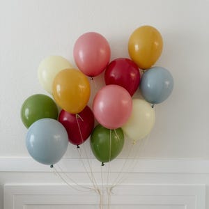 May include: A cluster of twelve colorful balloons in shades of pink, yellow, green, red, and blue. The balloons are tied together with string, creating a cheerful and festive arrangement. Perfect for a birthday or celebration.