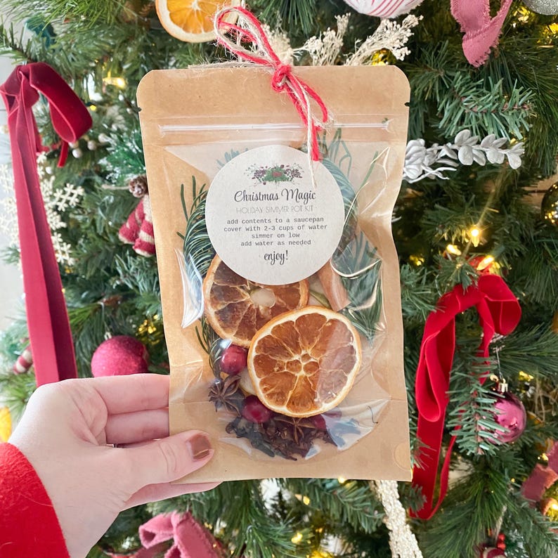 Puede incluir: Una bolsa de pl&aacute;stico transparente llena de rodajas de naranja secas, palitos de canela, an&iacute;s estrellado, clavos y ar&aacute;ndanos. La bolsa est&aacute; atada con un cordel rojo y blanco y tiene una etiqueta blanca que dice "Christmas Magic Holiday Simmer Kit" con instrucciones para agregar el contenido a una cacerola, cubrir con 2-3 tazas de agua, dejar hervir a fuego lento y agregar agua seg&uacute;n sea necesario. La etiqueta termina con "enjoy!"