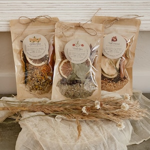 Peut inclure: Trois sachets transparents remplis de plantes et d'&eacute;pices s&eacute;ch&eacute;es, nou&eacute;s avec des n&oelig;uds en ficelle. Chaque sachet porte une &eacute;tiquette ronde avec du texte. Les sachets sont &eacute;tiquet&eacute;s "Golden Glow", "Forest Spice" et "Fall Harvest". Des fleurs et des herbes s&eacute;ch&eacute;es sont au premier plan.