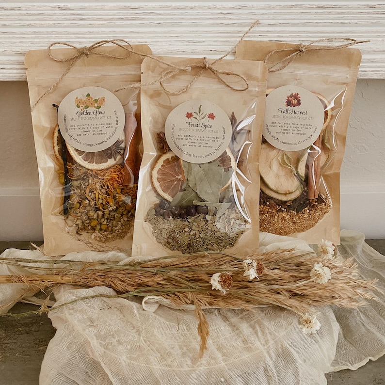Peut inclure: Trois sachets transparents remplis de plantes et d'&eacute;pices s&eacute;ch&eacute;es, nou&eacute;s avec des n&oelig;uds en ficelle. Chaque sachet porte une &eacute;tiquette ronde avec du texte. Les sachets sont &eacute;tiquet&eacute;s "Golden Glow", "Forest Spice" et "Fall Harvest". Des fleurs et des herbes s&eacute;ch&eacute;es sont au premier plan.